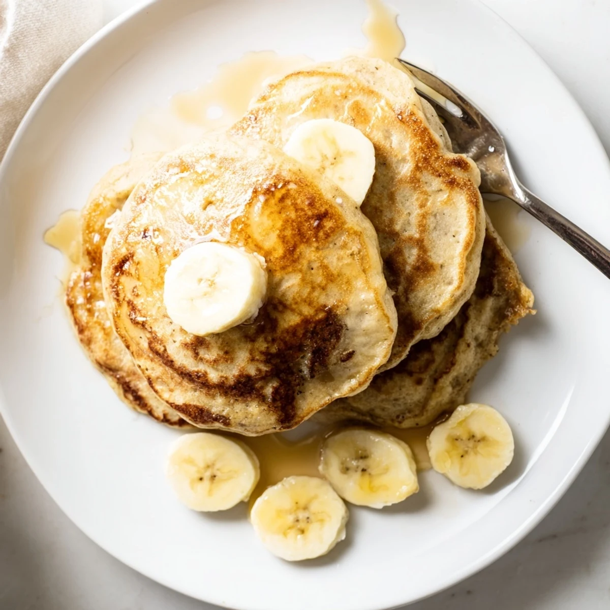 Golden stack of Greek yogurt banana pancakes drizzled with maple syrup and fresh banana slices on a white plate