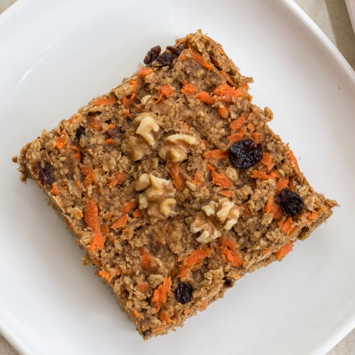 Moist flourless oatmeal carrot cake slice topped with chopped walnuts and raisins on a white plate